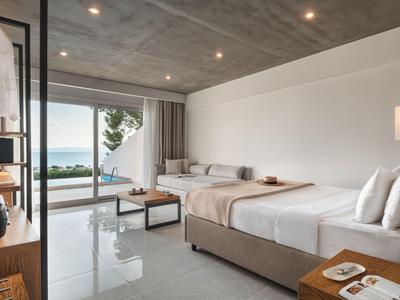 Modern hotel room with double bed, seating area, and sea view through large glass windows.