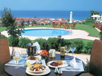 Tisch mit Essen vor Swimmingpool, dahinter Meer und Leuchtturm, Garten und Stühle umgeben den Pool.