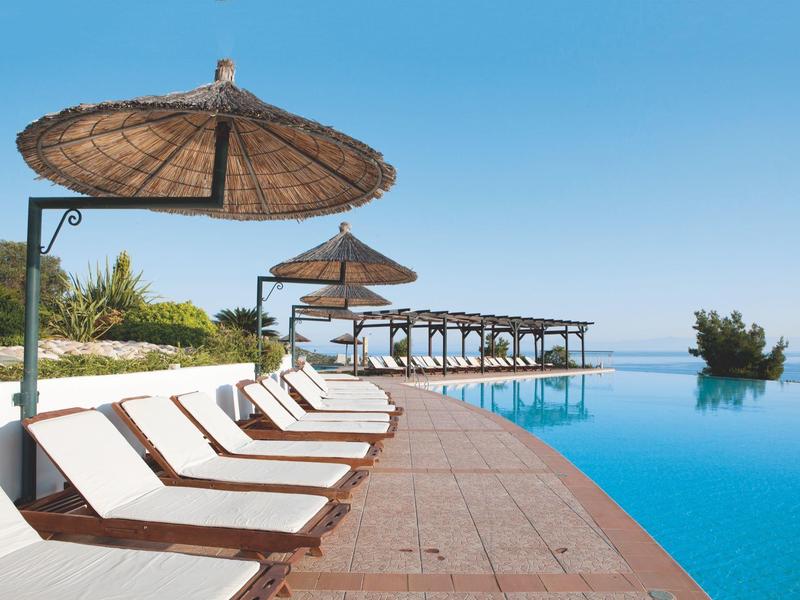 Infinity-Pool mit weißen Liegen, Strohdach-Sonnenschirmen und Blumen in klarem, blauem Himmel.