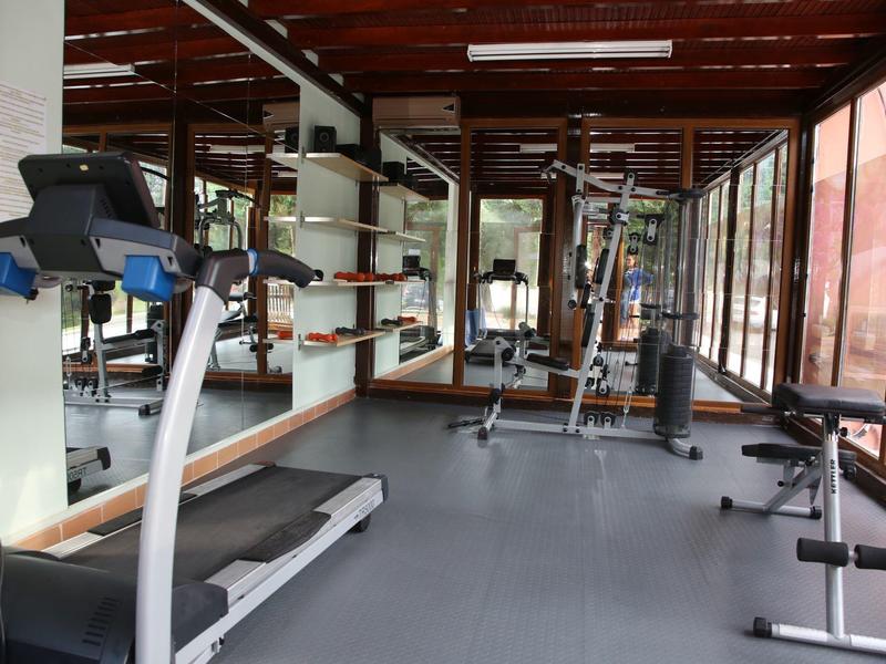 Fitnessraum mit Laufband, Hantelbank, großen Spiegeln und Glasfenstern an der Seite.