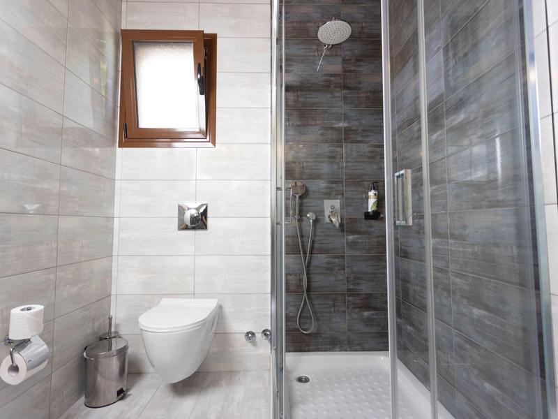 Kleines Badezimmer mit weißem WC, Duschkabine, grauen Fliesen und Holzfenster.