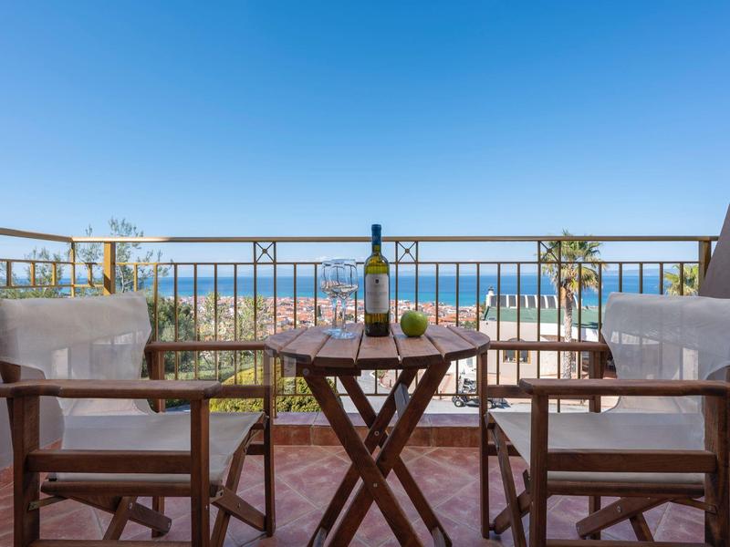 Balkon mit zwei Holzstühlen, kleinem Tisch, Pflanzen und Meerblick unter blauem Himmel.