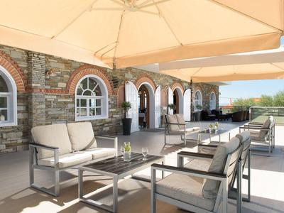 Terrassenmöbel mit grauen Polstern unter großen hellen Sonnenschirmen an Steinwand mit Rundfenstern.