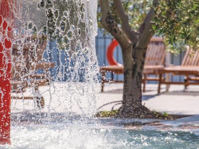 Wasserfontäne in einem Park mit Bäumen, Bänken und sonnigem, klarem Himmel.
