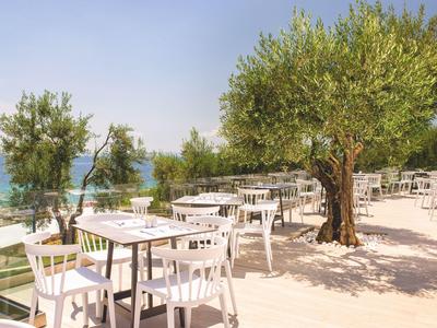 Terrasse mit weißen Tischen und Stühlen unter Olivenbäumen mit Meerblick bei klarem Himmel.