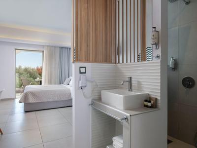 Moderner Schlafzimmerbereich mit weißem Bett, gegenüber Waschbecken in Bad mit Holzakzent und Duschkabine.