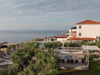 Hotelanlage mit Pool, Liegestühlen und mediterraner Vegetation am Meer unter bewölktem Himmel.