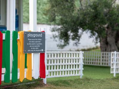 Bunter Kinderspielplatz mit Zaun und Hinweisschild neben weißem Gartenzaun.