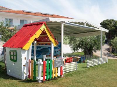 Kleines buntes Spielhaus auf einer grünen Rasenfläche neben einer überdachten Terrasse eines Hotels.