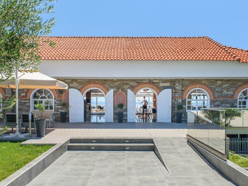 Eingang eines Hauses mit roten Ziegeldach, Steinmauern, drei Bögen und weißen Türen, umgeben von Grün.