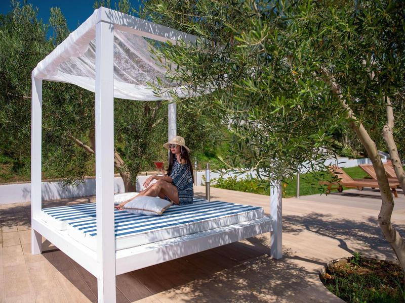 Weiße Gartenliege mit Baldachin unter Bäumen, Frau sitzt entspannt, blauer Himmel.