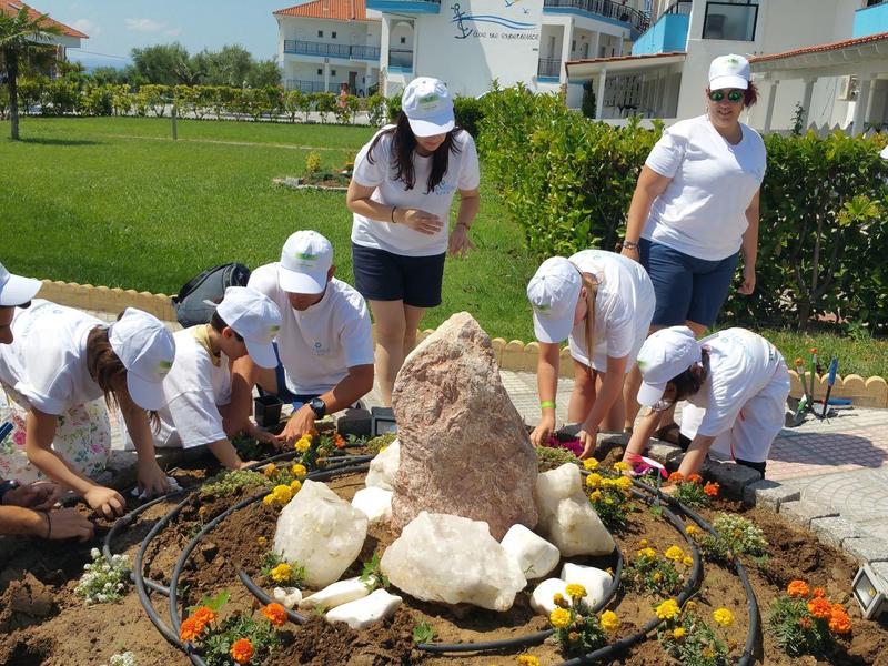 Kinder mit weißen Mützen arbeiten gemeinsam in einem Garten an einer Blumenpflanzung bei Sonnenschein.
