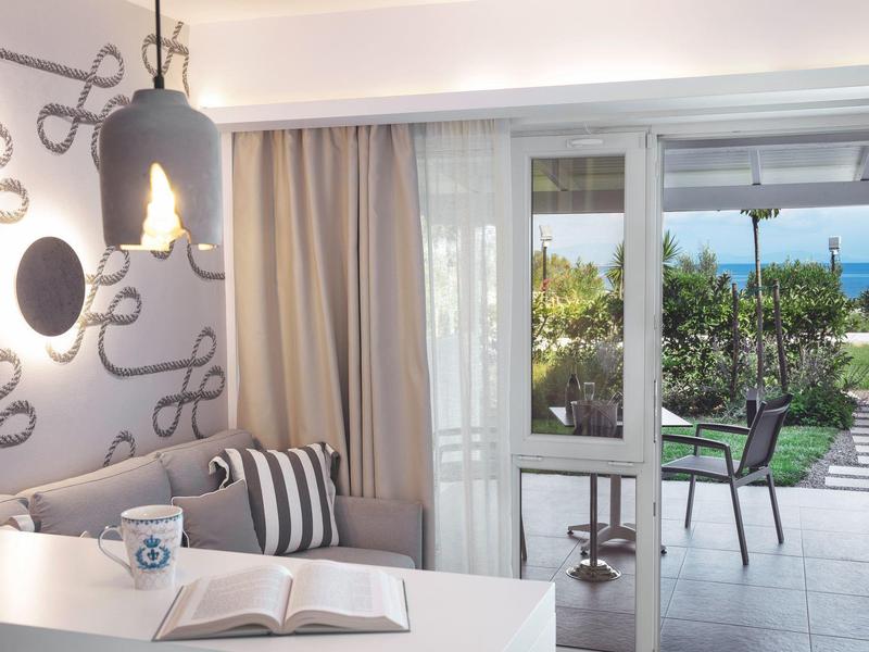 Helles Wohnzimmer mit grauem Sofa, Kissen, Tisch, aufgeschlagenem Buch und Blick auf Terrasse und Garten.