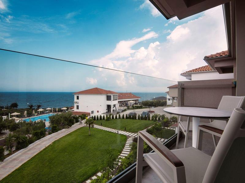 Balkon mit Stühlen und Tisch, Blick auf grünen Garten, weiße Gebäude und blauen Himmel am Meer.