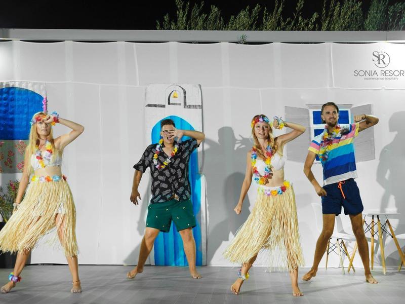 Menschen tanzen bunt gekleidet vor heller Wand mit Schirmlogo bei Nacht.