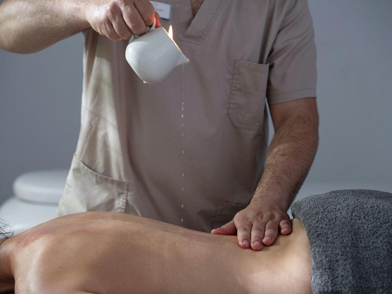 Mann gießt Wasser aus einer weißen Tasse auf den nackten Rücken einer liegenden Person.