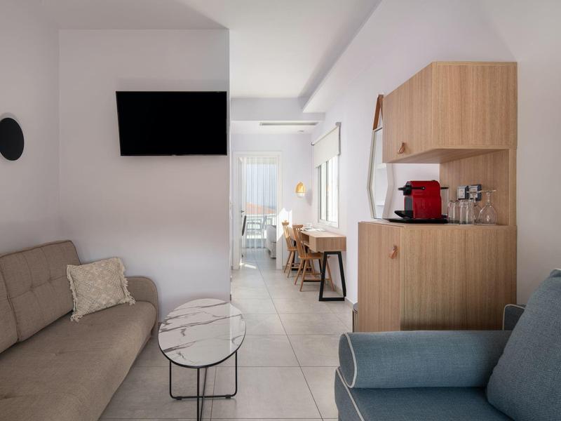 Kleines, helles Wohnzimmer mit Sofa, Sessel, Wand-TV, kleinem Tisch und Küchenzeile aus Holz.