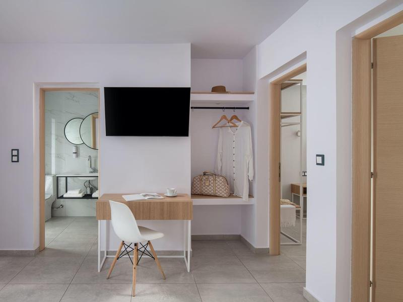 Moderner Hotelraum mit Schreibtisch, Fernseher und offener Garderobe mit Taschen.