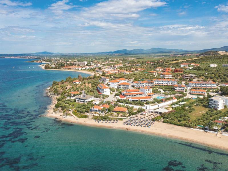 Küstenhotel mit Sandstrand, Poolbereich und umliegender mediterraner Landschaft unter blauem Himmel.