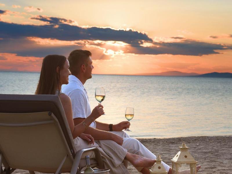 Paar sitzt entspannt auf Strandliegen, genießt Sonnenuntergang mit Weingläsern am Strand.
