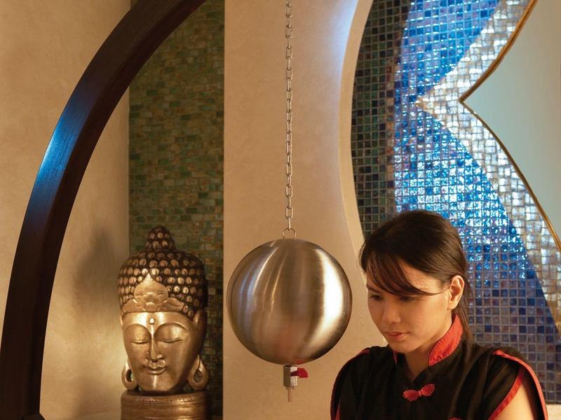 Eine Frau massiert im Spa-Bereich eines Hotels sanft den Kopf eines Gastes vor einem Buddha-Bild.