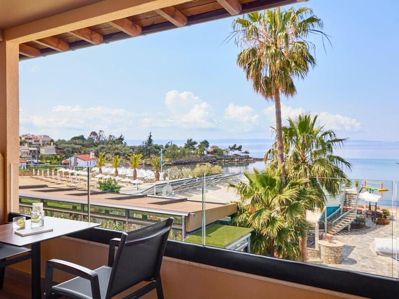 Balkon mit Tisch und Stühlen, Blick auf Palmen, Pool und Meer am Strand.