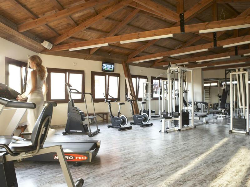 Helles Fitnessstudio mit Holzdecke, zahlreichen Trainingsgeräten und einer Frau auf dem Laufband.