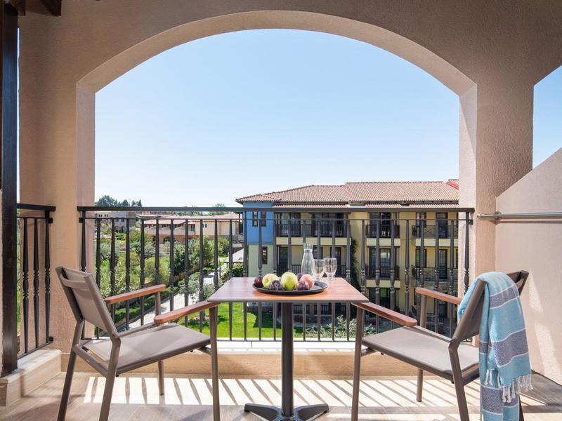Balkon mit Tisch und zwei Stühlen mit Blick auf Gebäude und Grünflächen bei klarem Himmel.