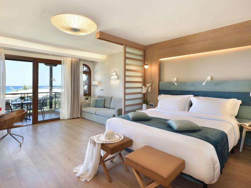 Helles Hotelzimmer mit Doppelbett, Sitzbereich und Balkon mit Meerblick.