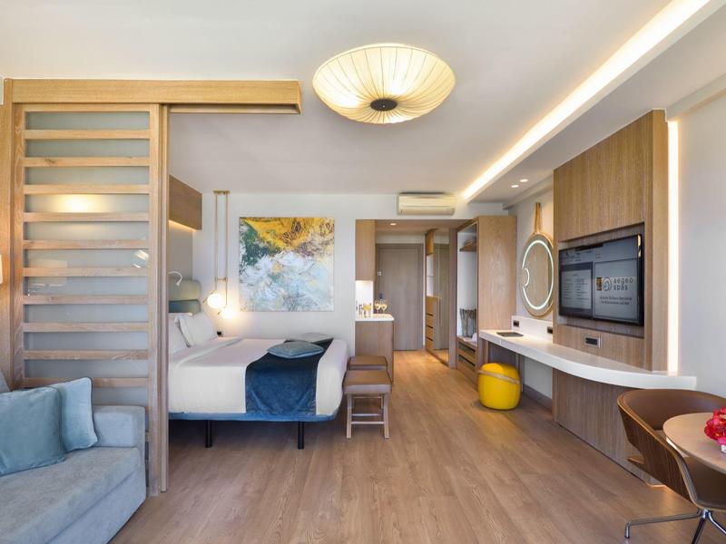 Modern eingerichtetes Hotelzimmer mit Schlafzimmer, Sitzbereich und Essplatz in warmen Farben.