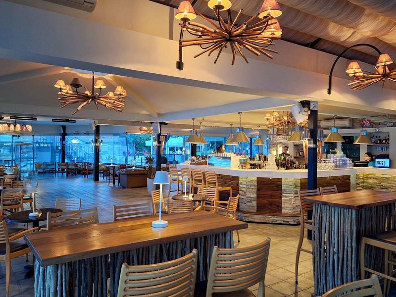 Modernes Restaurant mit Holzmöbeln und großer Fensterfront mit Blick auf das Meer bei Sonnenuntergang.