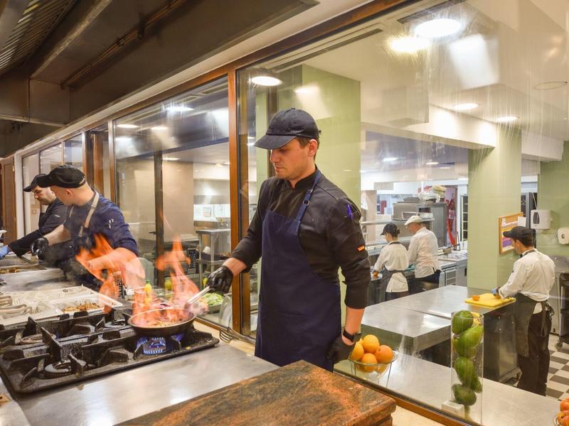 Zwei Köche bereiten in einer professionellen Restaurantküche Speisen mit offener Flamme zu.