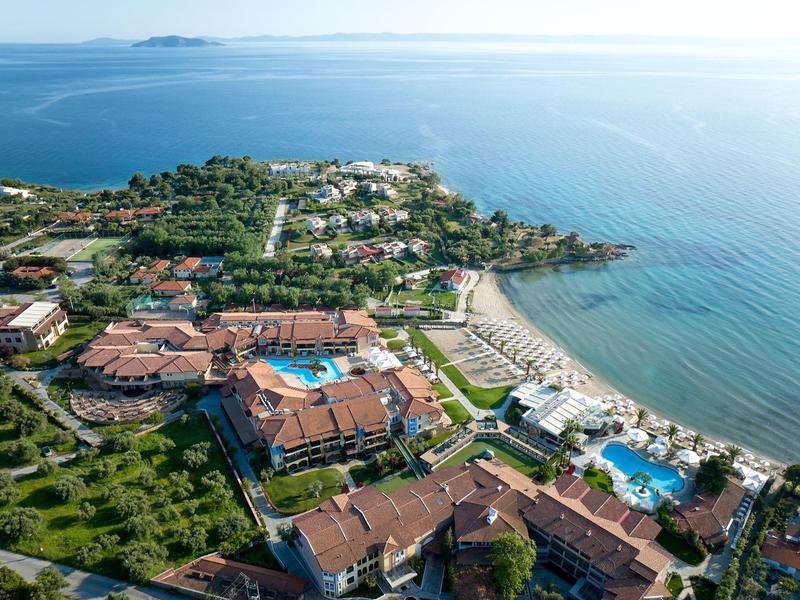 Luftaufnahme eines Küstenhotels mit Pools, Gartenanlage und Strand am blauen Meer.