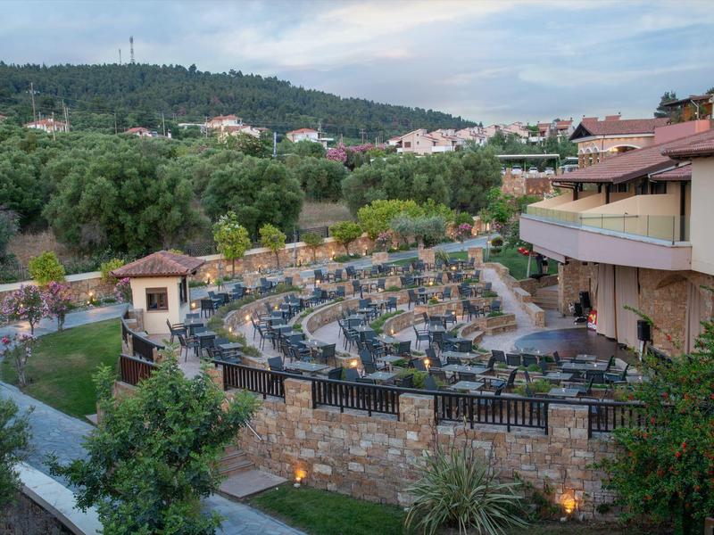 Terrassenförmiger Außenbereich eines Hotels mit Tischen und Stühlen, umgeben von Bäumen und Hügeln.