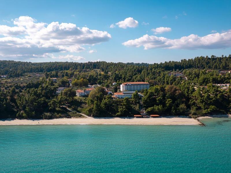 Hotel u pláže obklopený hustým lesem a tyrkysovým mořem pod částečně oblačnou oblohou.