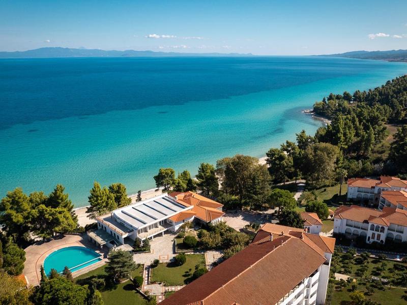 Pobřežní resort s bazénem, obklopený stromy a budovami u modrého moře pod jasnou oblohou.