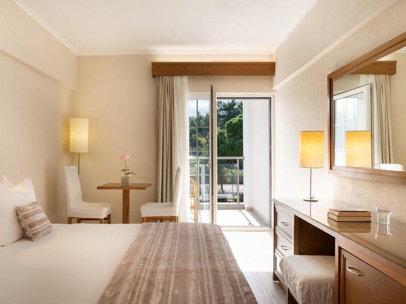 Světý hotelový pokoj s postelí, stolem, židlí a balkonem s bujnou zelení venku.