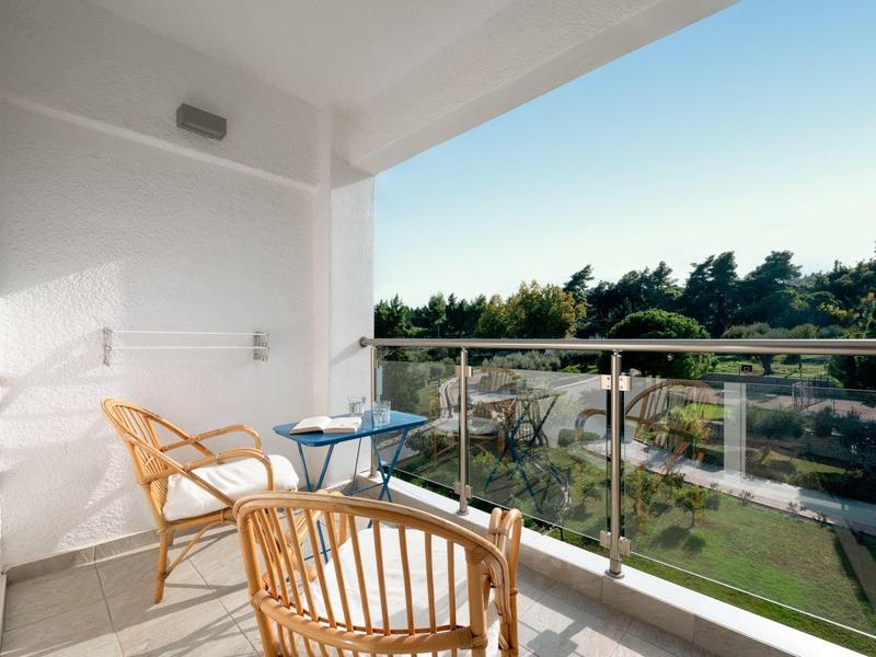 Balkon s ratanovými židlemi a skleněným stolem s výhledem na zelenou zahradu pod jasnou oblohou.