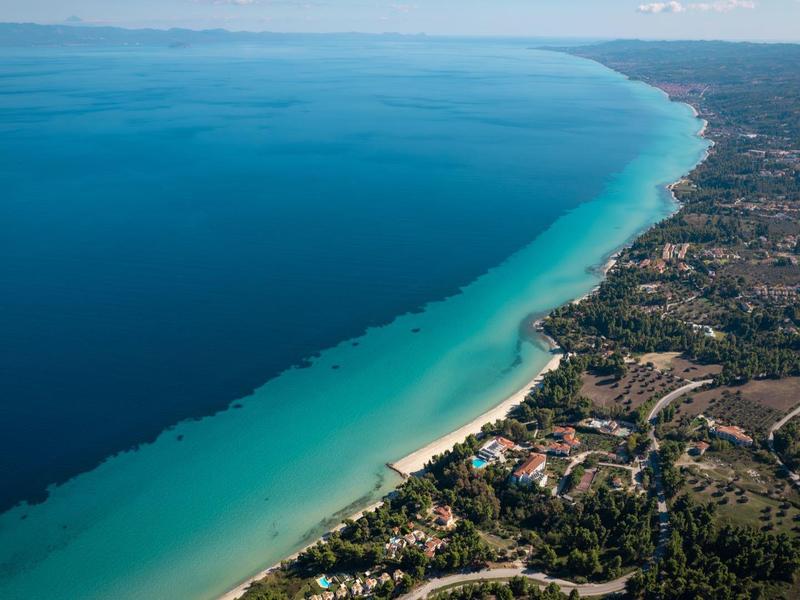 Letecký pohled na dlouhou písečnou pláž s tyrkysovou vodou a zalesněnou krajinou.
