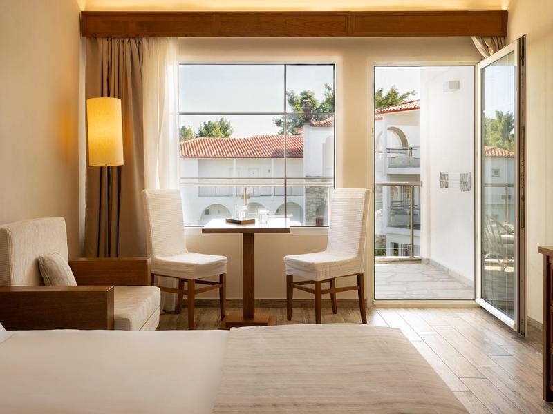 Pohodlný hotelový pokoj s postelí, křeslem, stolem, židlemi, lampou a otevřenými dveřmi na balkon se slunečným výhledem.