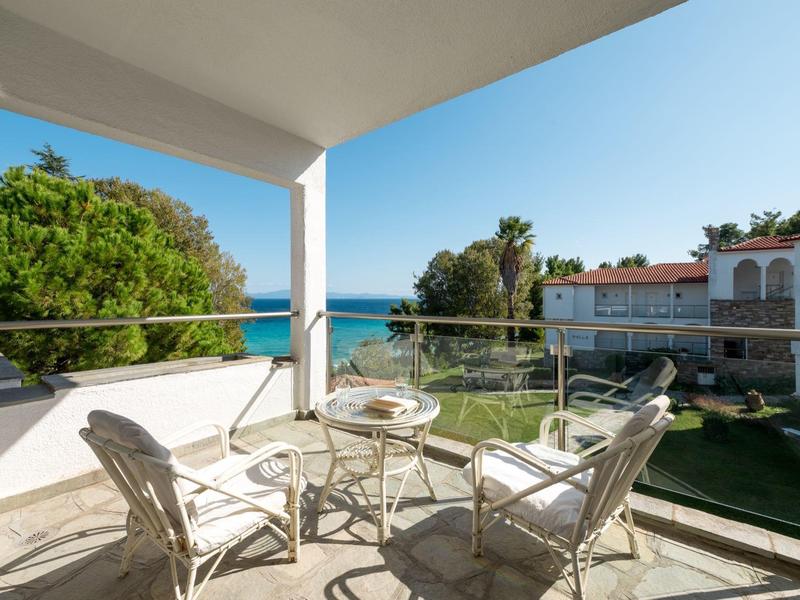 Balkon s skleněným stolem a židlemi s výhledem na zahradu a moře pod modrou oblohou.
