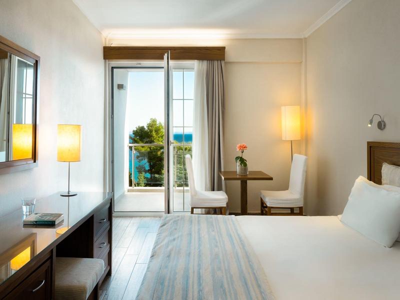 Světle zařízený hotelový pokoj s manželskou postelí, dvěma židlemi u balkonu a teplým osvětlením.