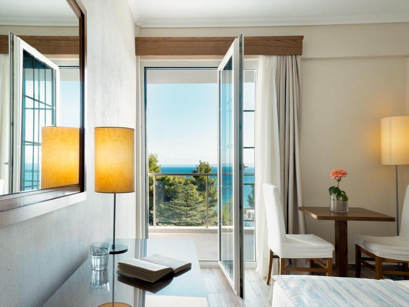 Hotelový pokoj s otevřenými balkónovými dveřmi, výhledem na moře, posezením a teplým osvětlením.