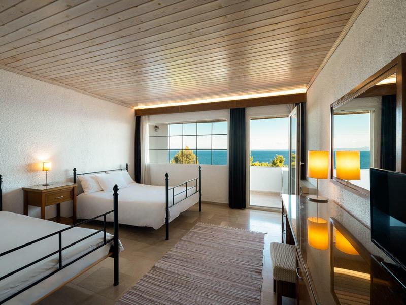 Prostorný hotelový pokoj se dvěma postelemi, dřevěným stropem a balkonem s výhledem na moře.