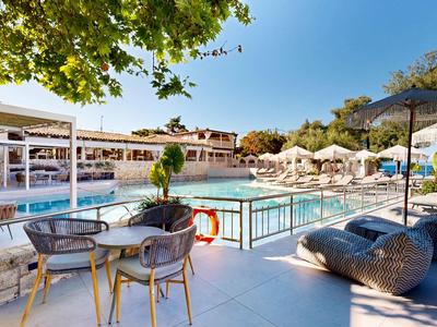 Area piscina esterna moderna con lettini e tavoli in un hotel per vacanze sotto un cielo blu.