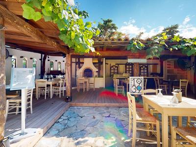 Freiluftrestaurant mit hellem Holzmobiliar, bunten Wanddekorationen und grünen Pflanzen unter blauem Himmel.