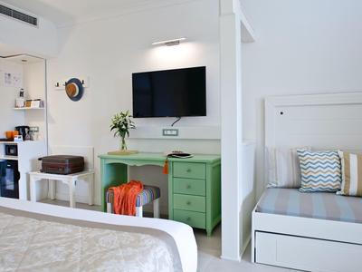 Bright hotel room with TV, green desk, bed, and bench with cushions.