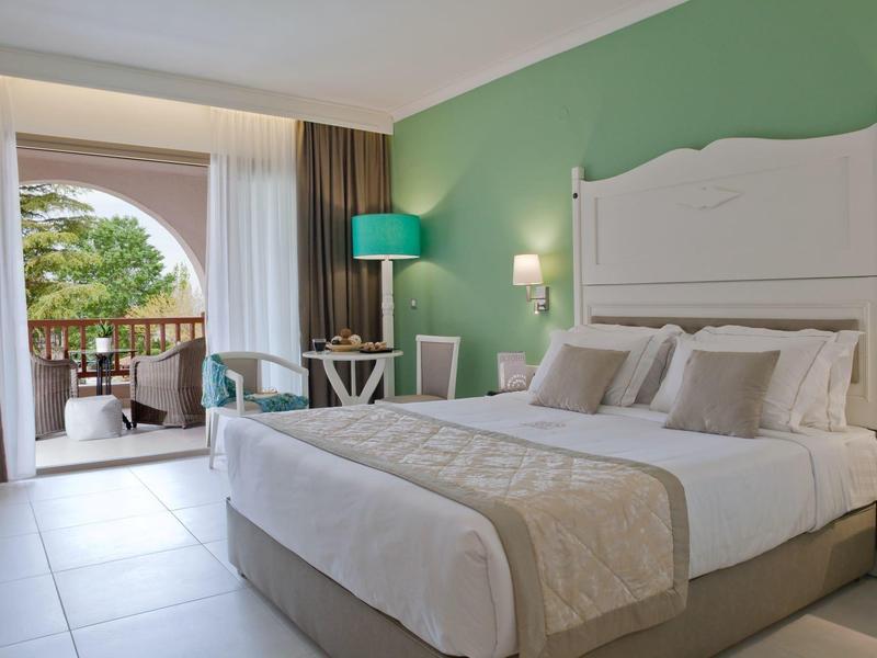 Bright hotel room with double bed, green accent wall, and balcony overlooking garden.