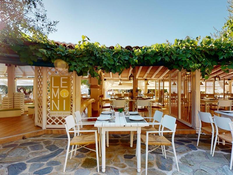 Outdoor restaurant with tables and chairs under a green wooden pergola on a clear sky day.