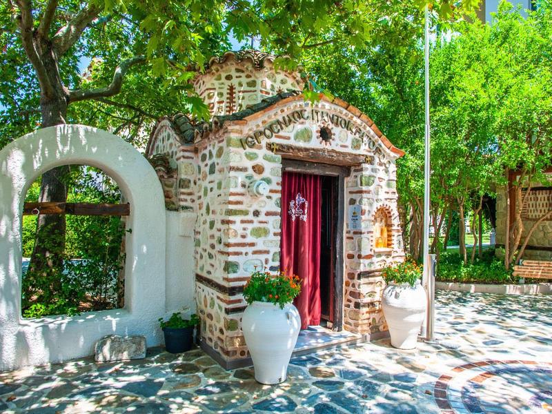 Small stone chapel with red door and floral decorations under green trees.